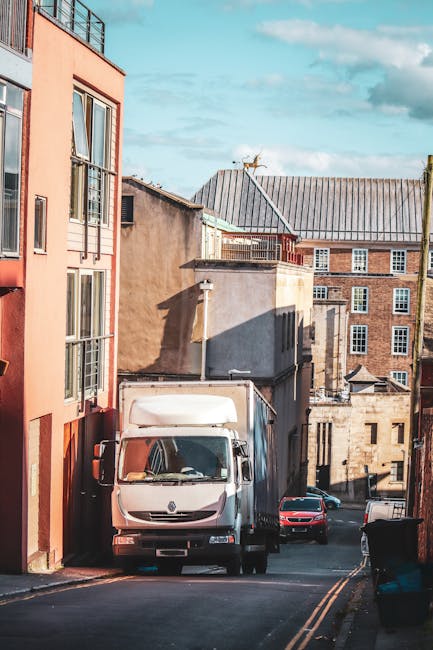 Wide-angle view of a city street featuring several historic and modern buildings with varied architectural styles, including brick facades, arched windows, and decorative details. A grey medium-sized