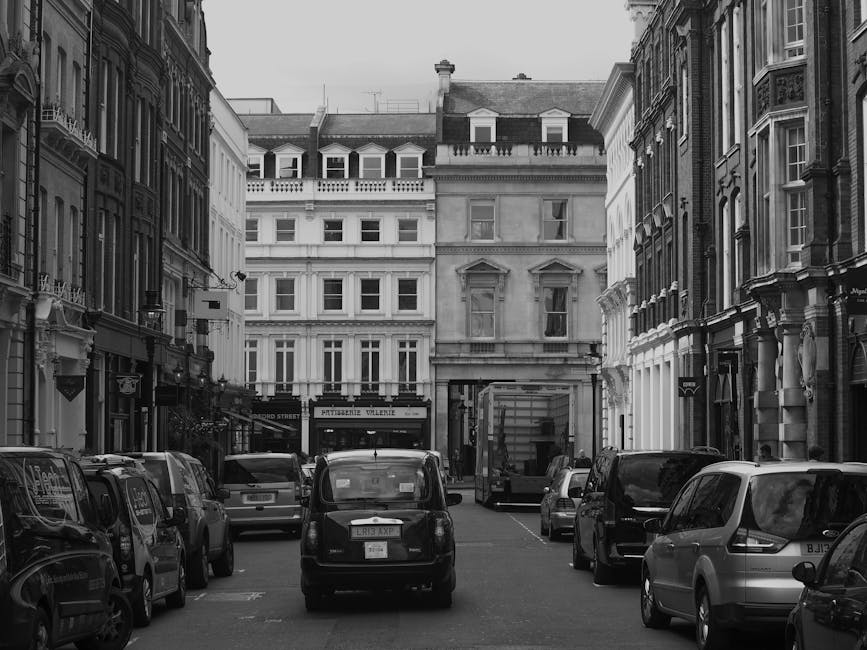 A grey commercial van parked on the street in front of a historic brick building with large arched windows and stone detailing around the entrance. The van is positioned parallel to the pavement, part